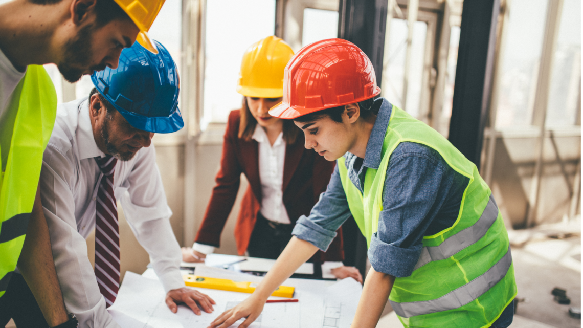 A hispanic woman, a white woman, and two hispanic men look over architectural plans. All are wearing hard hats.
