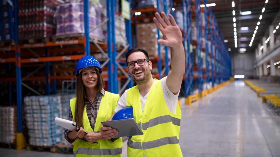 One male and one female manufacturing professional smiling and waving in a warehouse